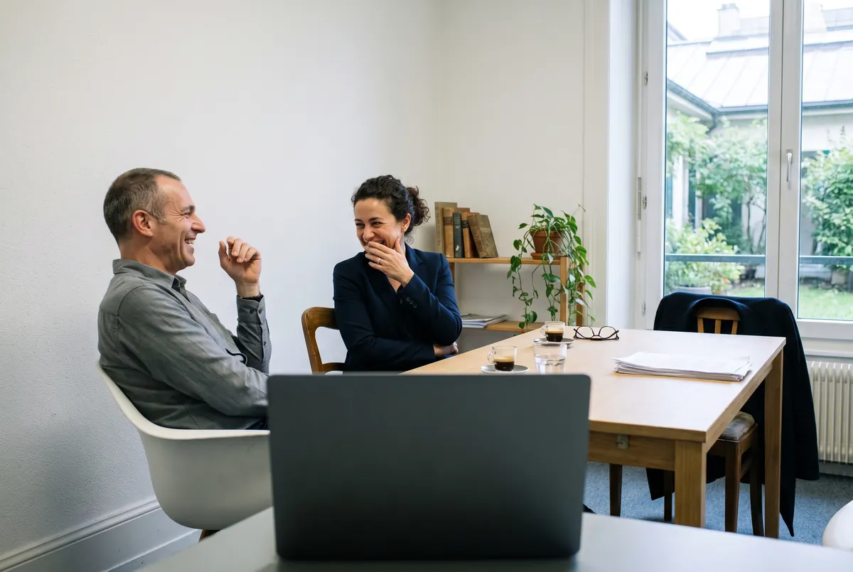 Un formateur et une élève rient ensemble autour d'une table de travail, deux tasses d'espresso posées à côté d'un ordinateur portable